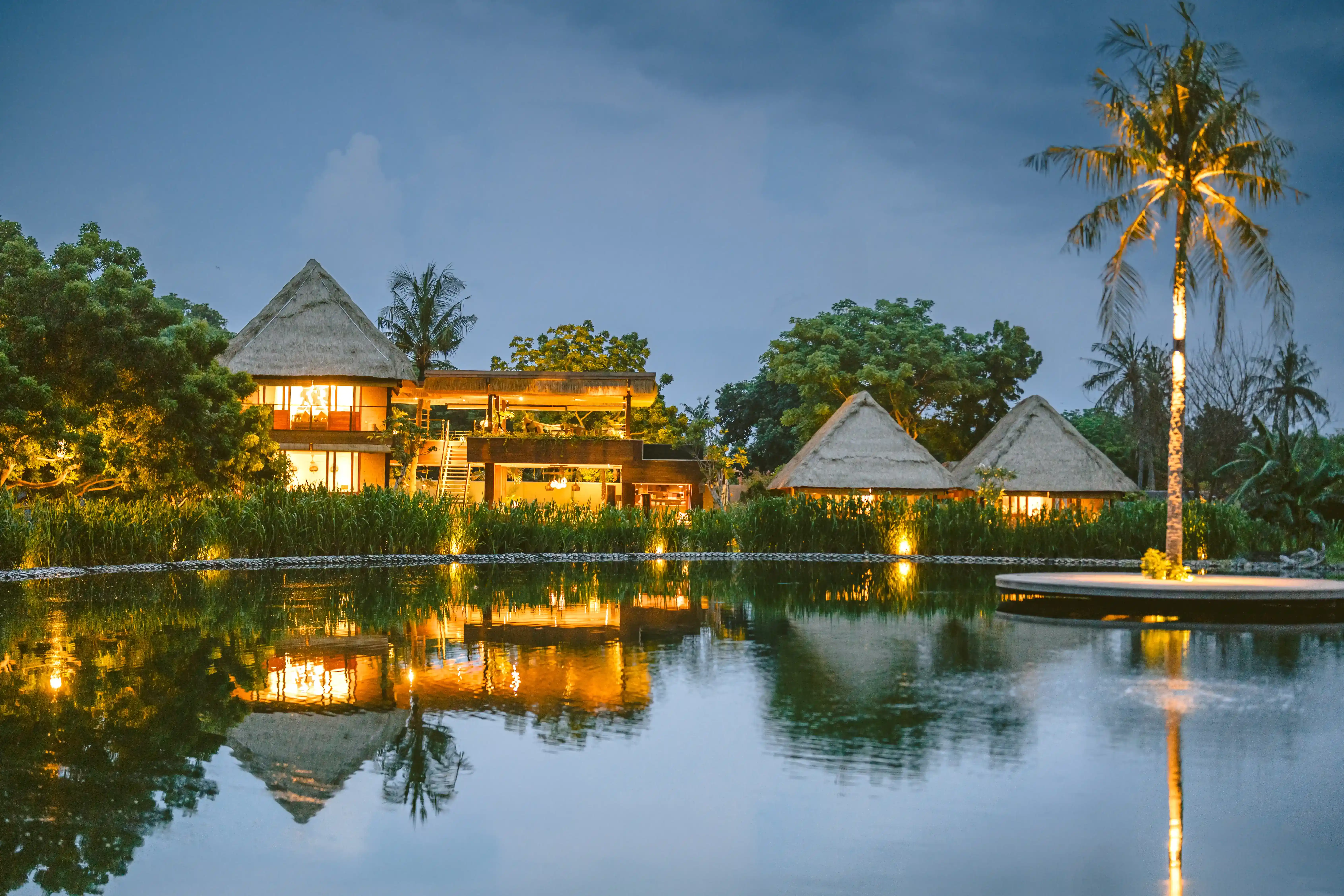 Twilight view of a two-story luxury villa featuring a long illuminated private pool and traditional thatched architecture.