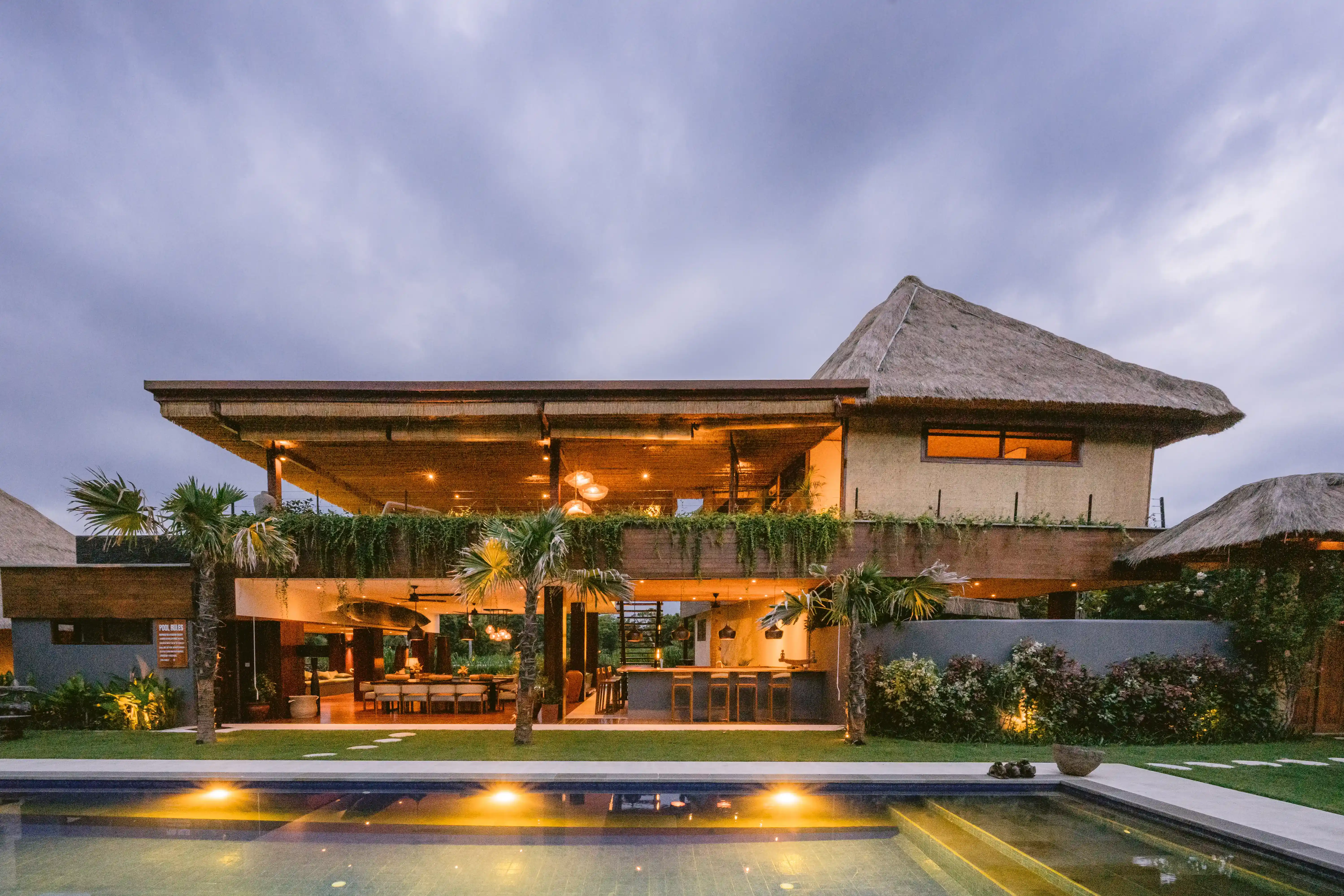 Twilight view of a two-story luxury villa featuring a long illuminated private pool and traditional thatched architecture.