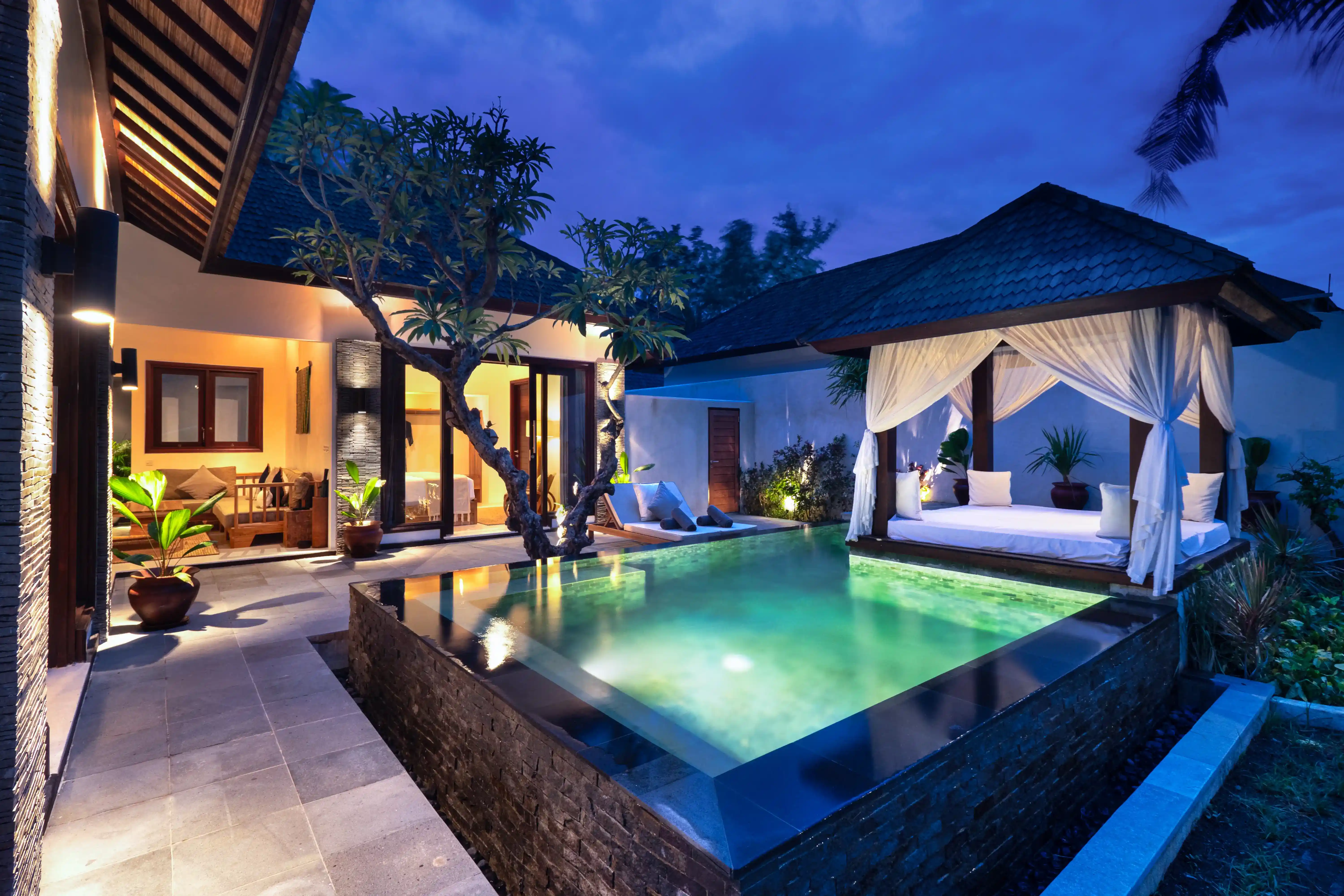 Open doorway view into the 2 Bedroom Pool Villa, showing a stylish wooden chair and modern floor lamp, with a stone accent wall outside
 hover