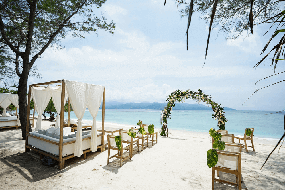 Wedding setup at Pondok Santi Estate, Gili Trawangan, with arch, decorated chairs on white sand beach, ocean, and Lombok in the background.