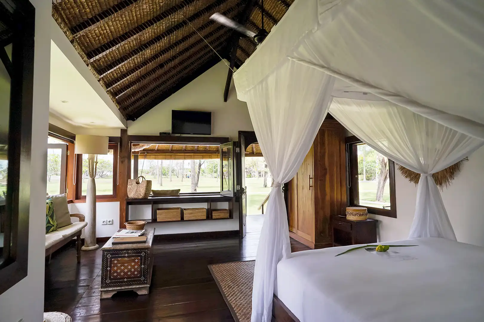 Interior view of the Beachside Bungalow queen-size bed with tropical pillows, looking out through the open wooden door to the private garden hover