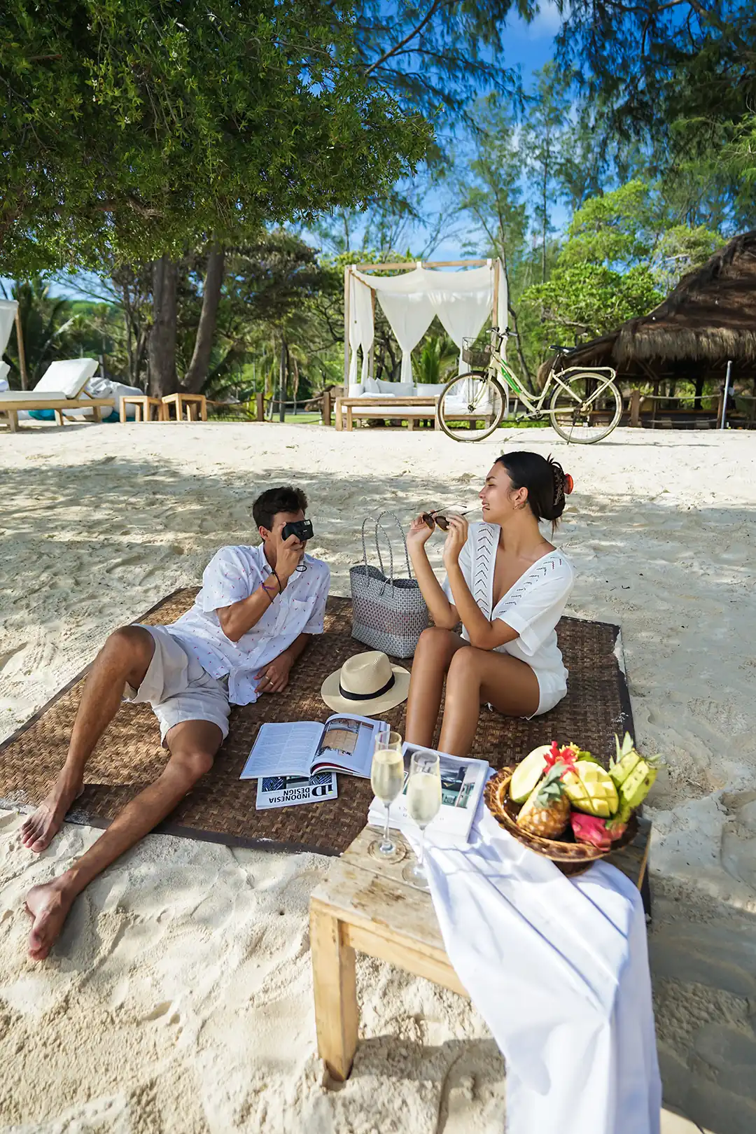 A couple enjoying a private beach picnic with fresh fruit and champagne, set on a mat with a daybed and bike visible in the background