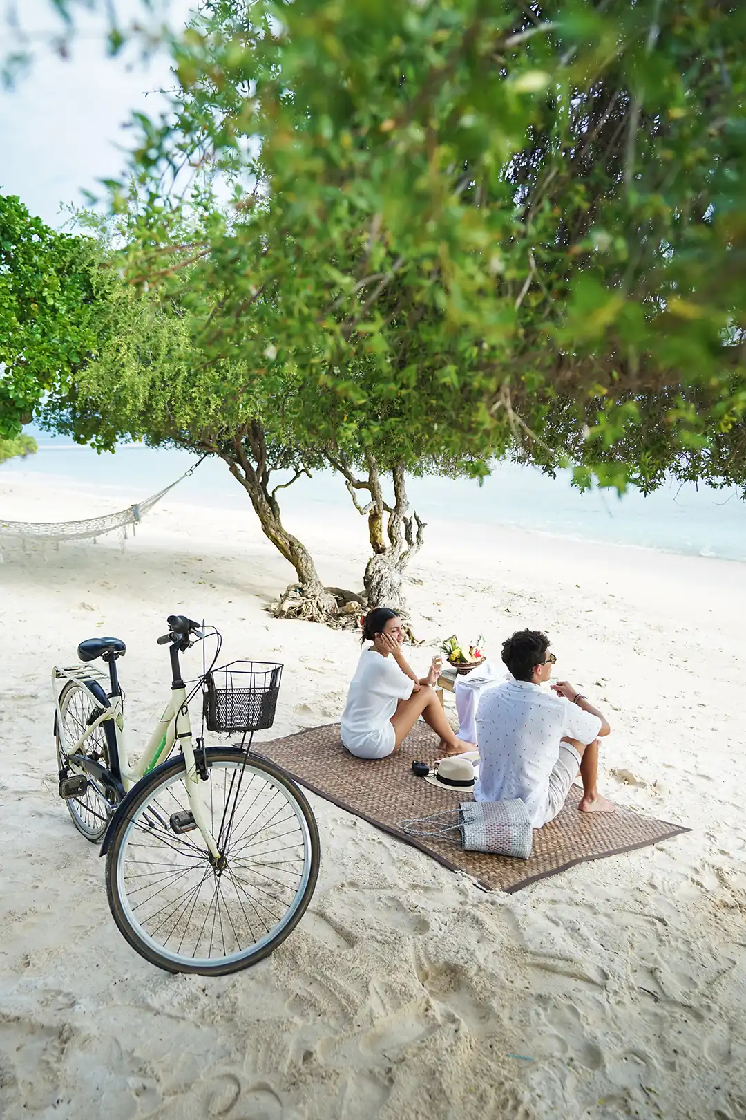 A couple enjoying a private beach picnic with fresh fruit and champagne, set on a mat with a daybed and bike visible in the background hover