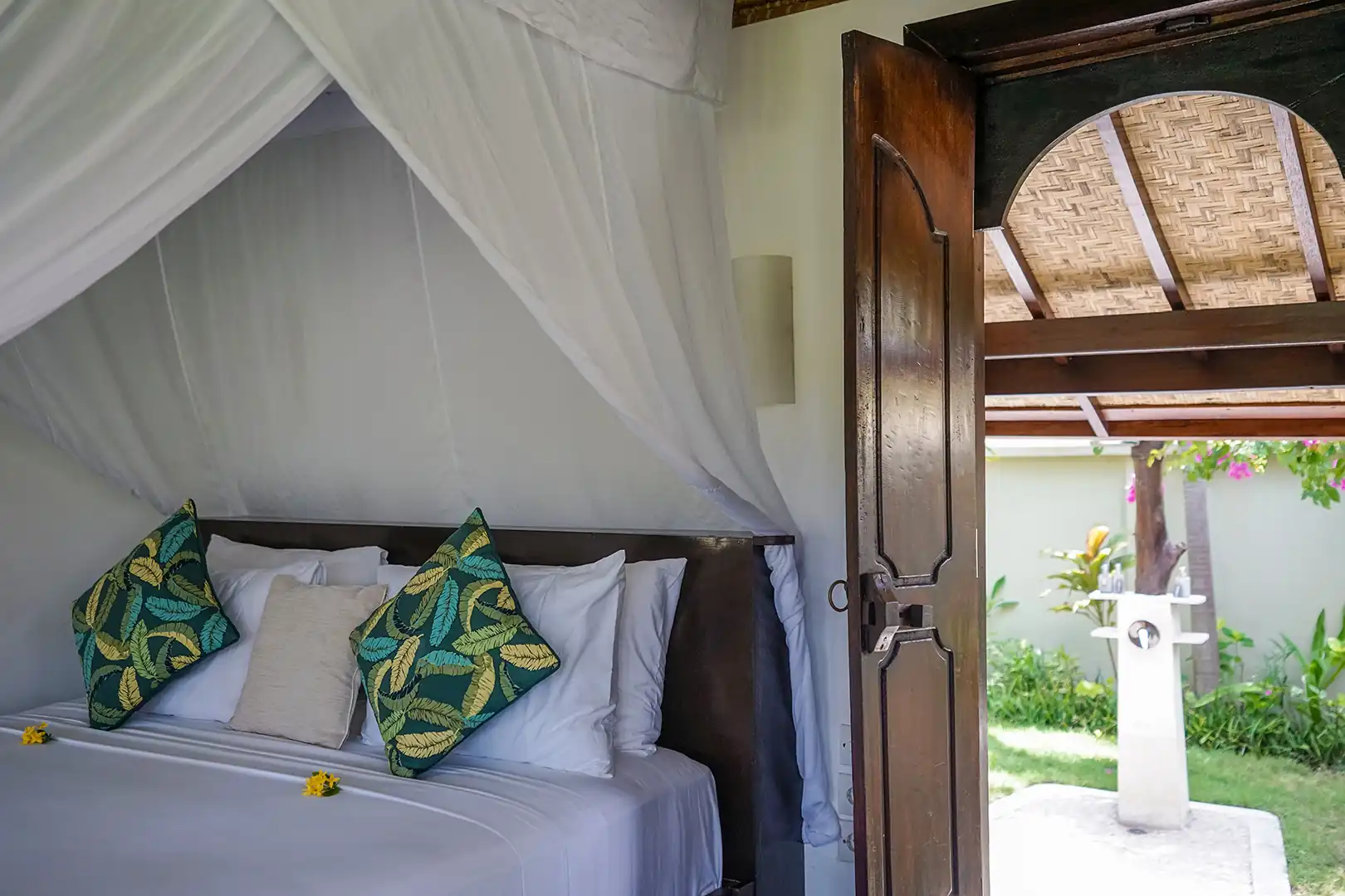 Interior view of the Beachside Bungalow queen-size bed with tropical pillows, looking out through the open wooden door to the private garden