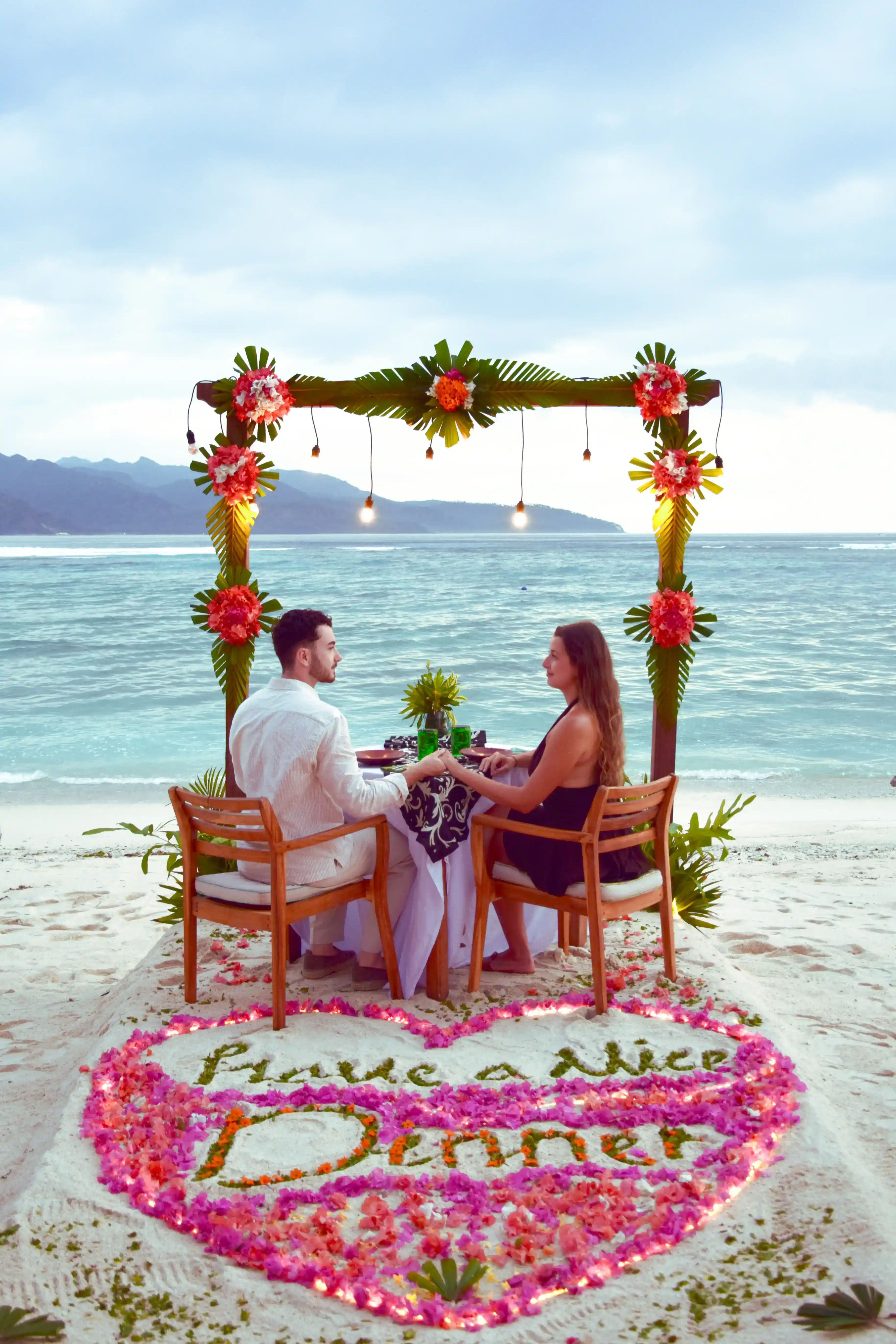 A couple sitting at a beautifully set table on the beach under a decorated arch enjoying a Romantic Dinner
