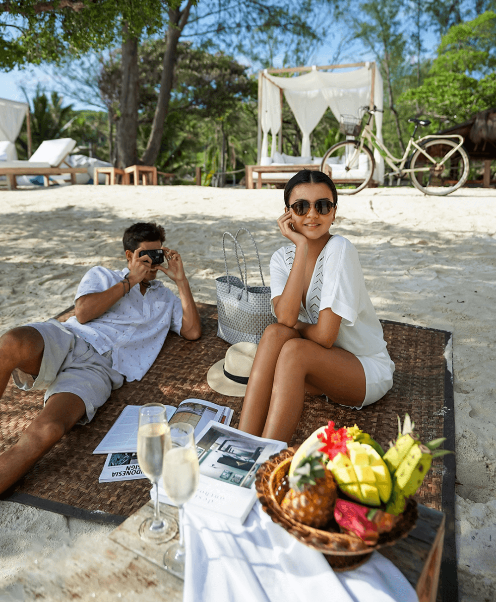 Man taking a picture of woman during a beach picnic with fruits at Pondok Santi Estate, Gili Trawangan, with a beach cabana in the background.