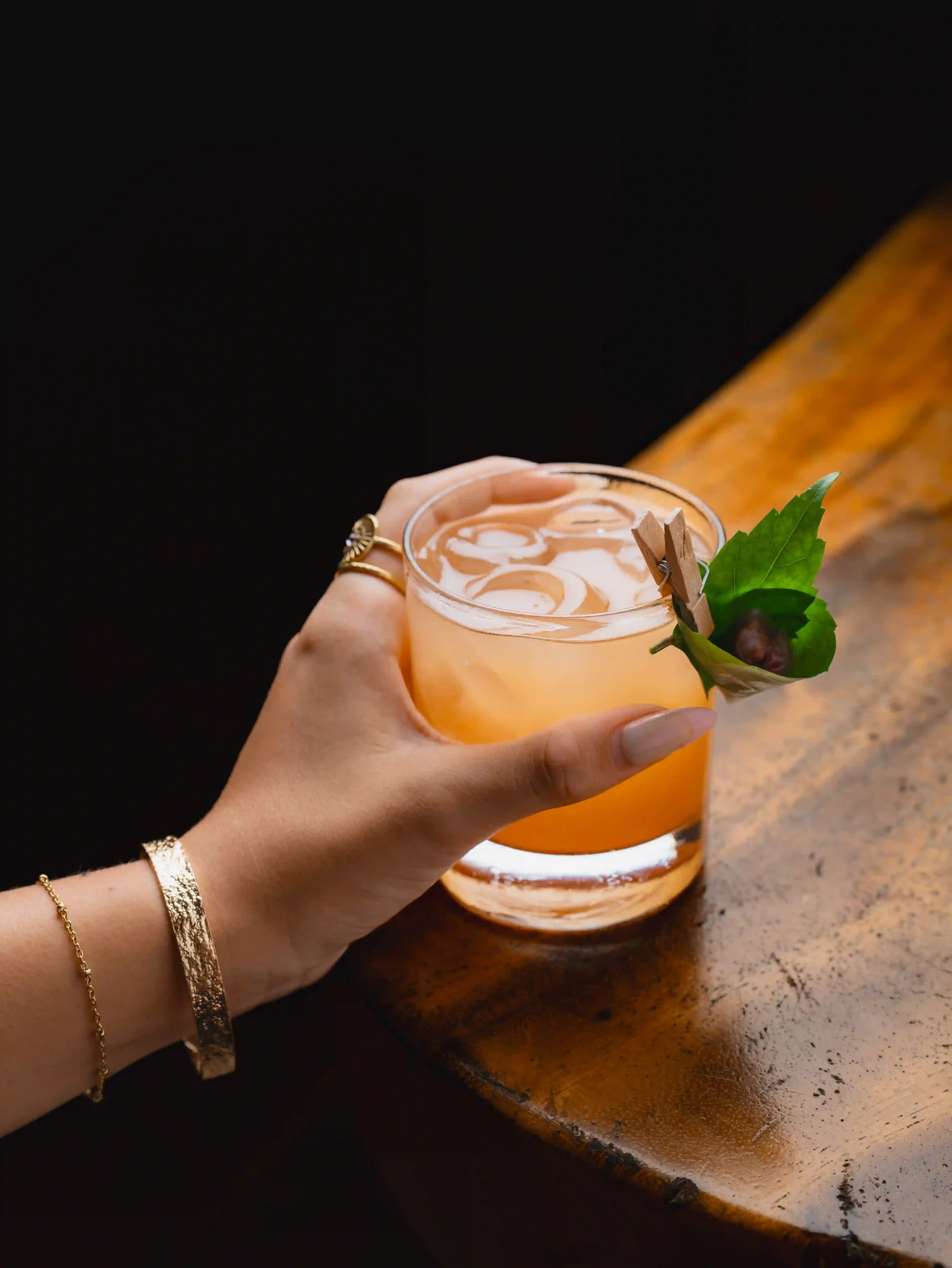A hand wearing a gold bracelet holding an orange cocktail with ice and a garnish, resting on a wooden bar surface during Happy Hour