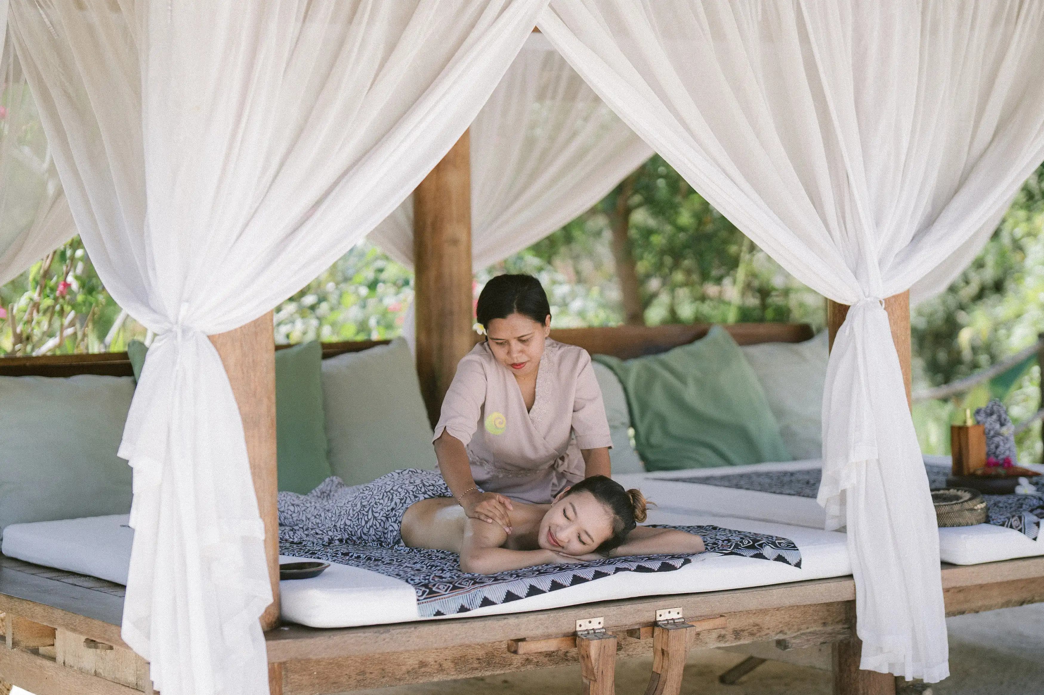 A guest receiving an in-room spa massage under a sheer white canopy on an outdoor daybed, performed by a professionally trained therapist