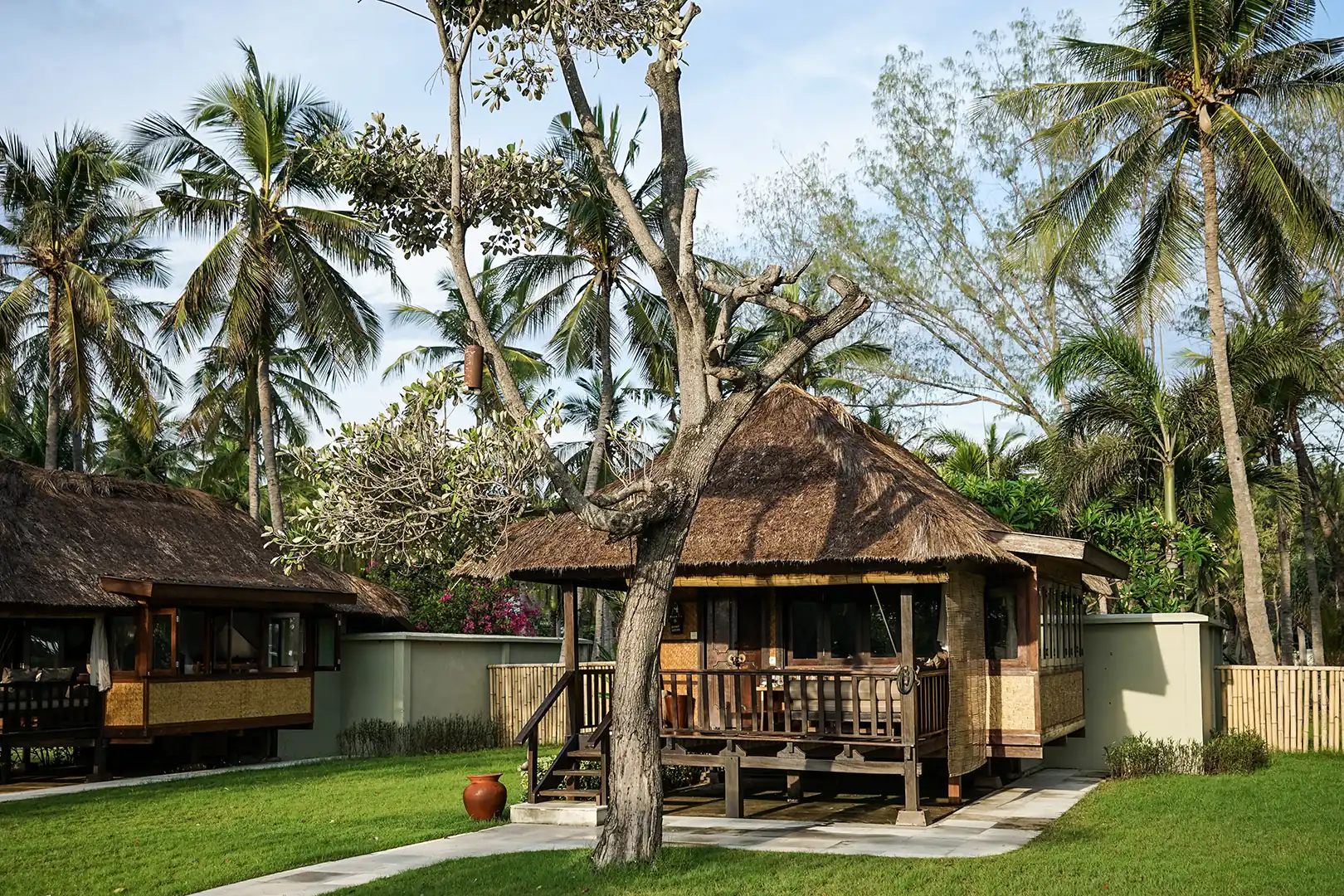 Exterior view of the Palm View Bungalow, showing the thatched roof, private wooden terrace, and palm trees surrounding the building