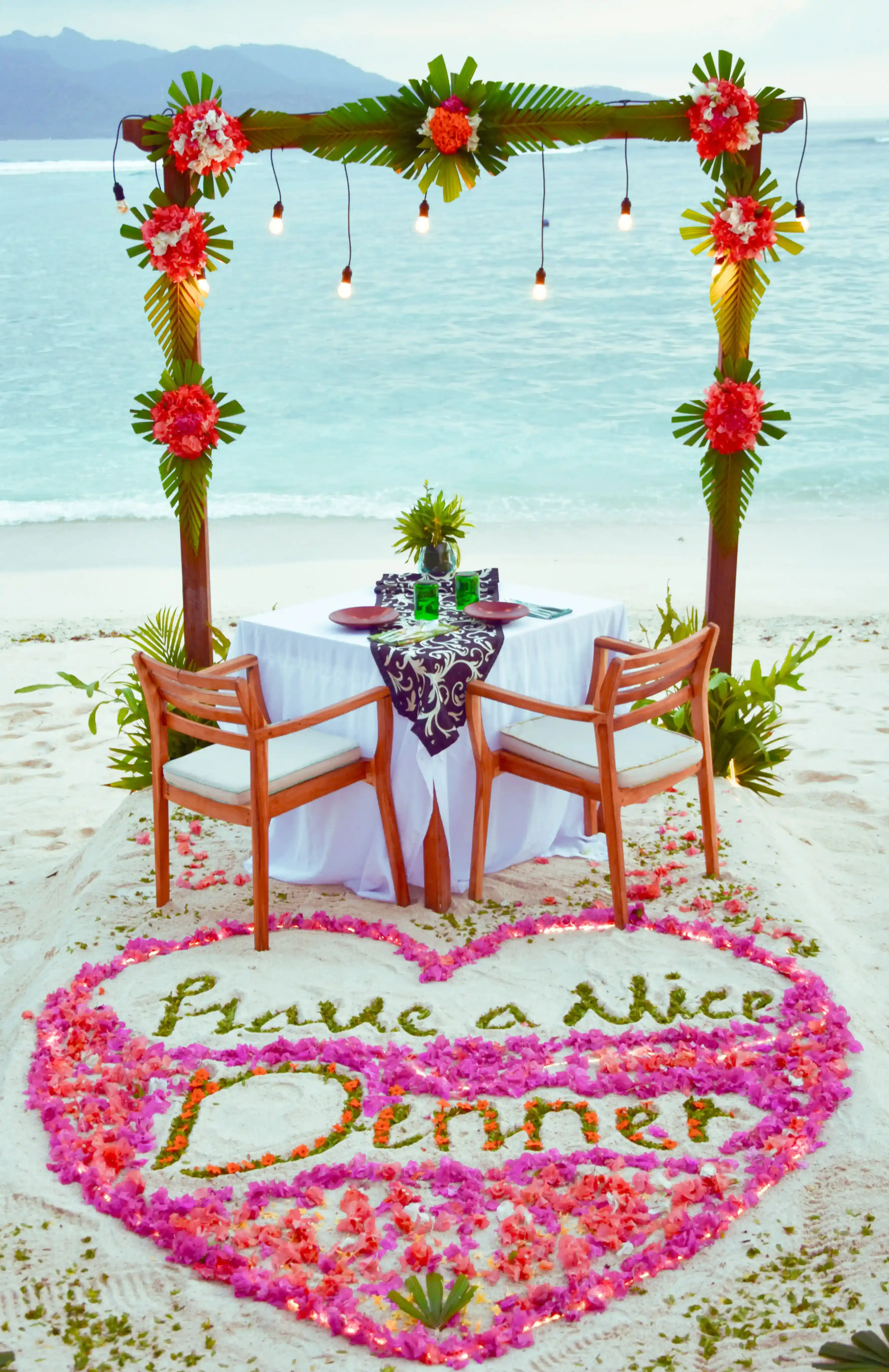 A couple sitting at a beautifully set table on the beach under a decorated arch enjoying a Romantic Dinner
 hover
