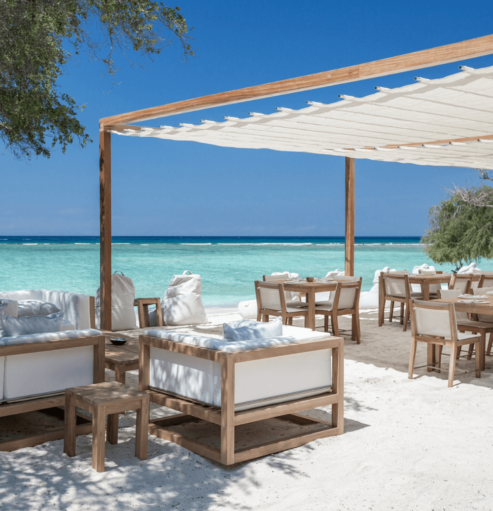 Santi Beach restaurant at Pondok Santi Estate, with cozy sofas and dining tables set on white sand beach overlooking clear waters.