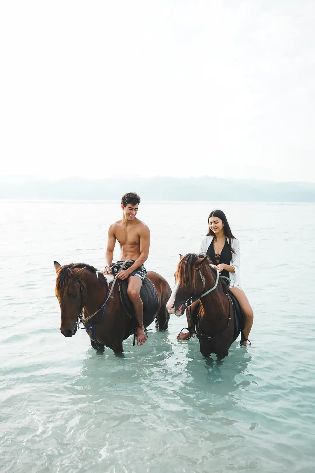 A person on horseback walking into the ocean at sunrise, with a guide leading, and large mountains visible in the distance across the water hover