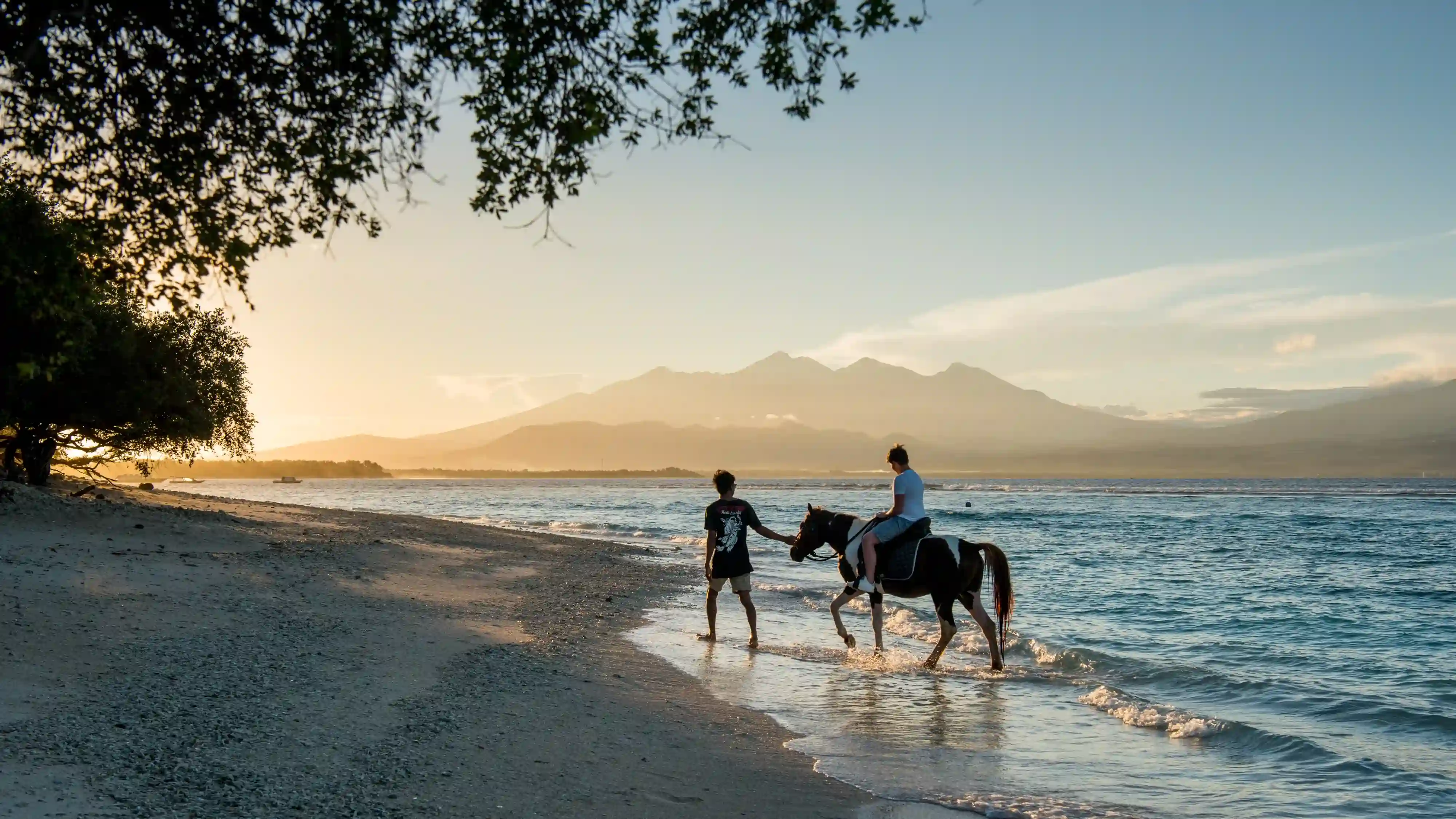 A person on horseback walking into the ocean at sunrise, with a guide leading, and large mountains visible in the distance across the water