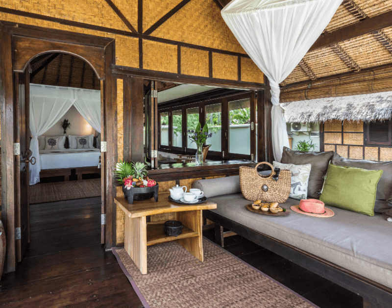 Terrace of Beachside Bungalow at Pondok Santi Estate, Gili Trawangan, with comfortable sofa, table with fruits, and view into the bungalow through open windows and door.