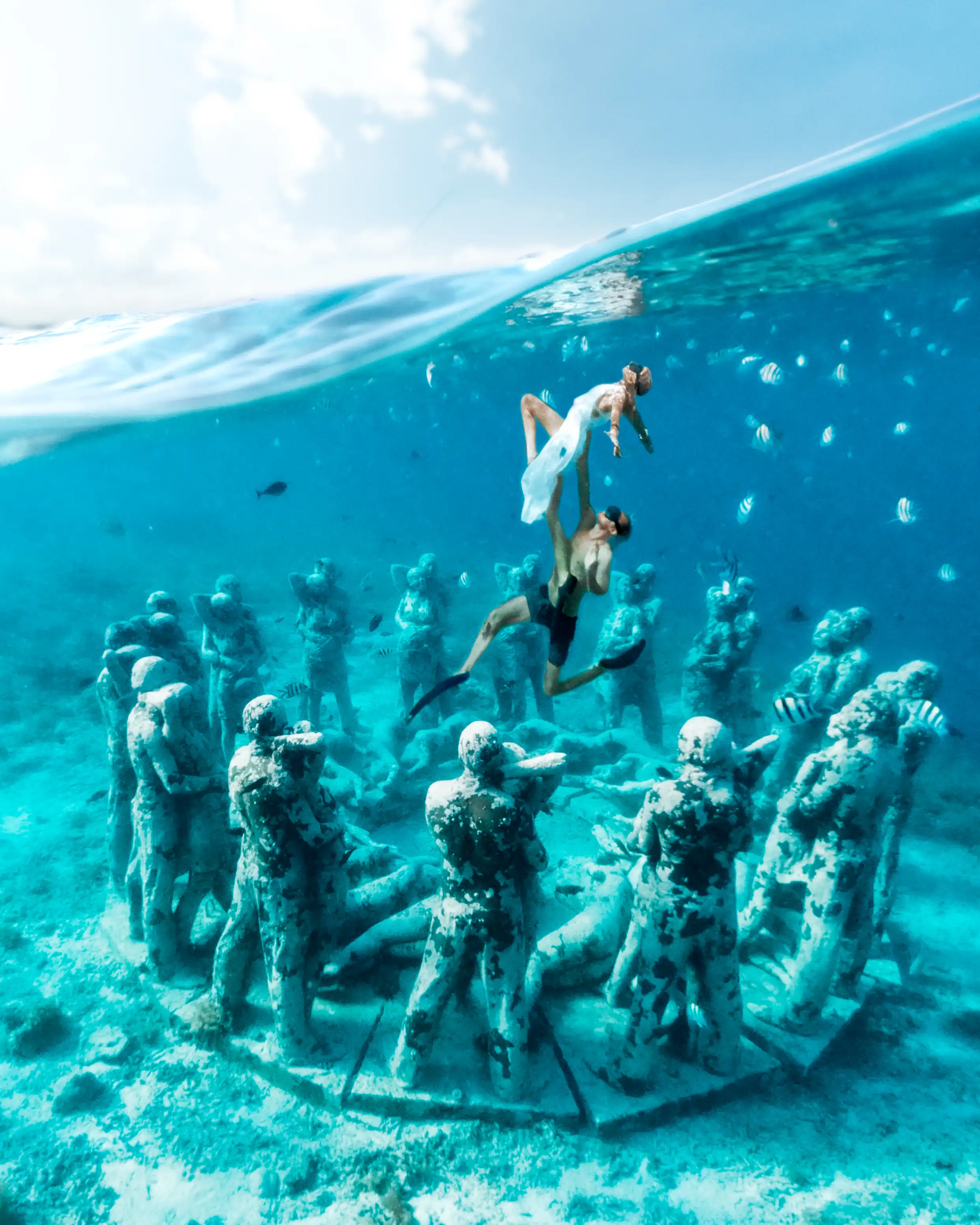 A couple swimming above the famous underwater statue circle, half-submerged photo with clear blue water and fish, representing complimentary Resort Activities