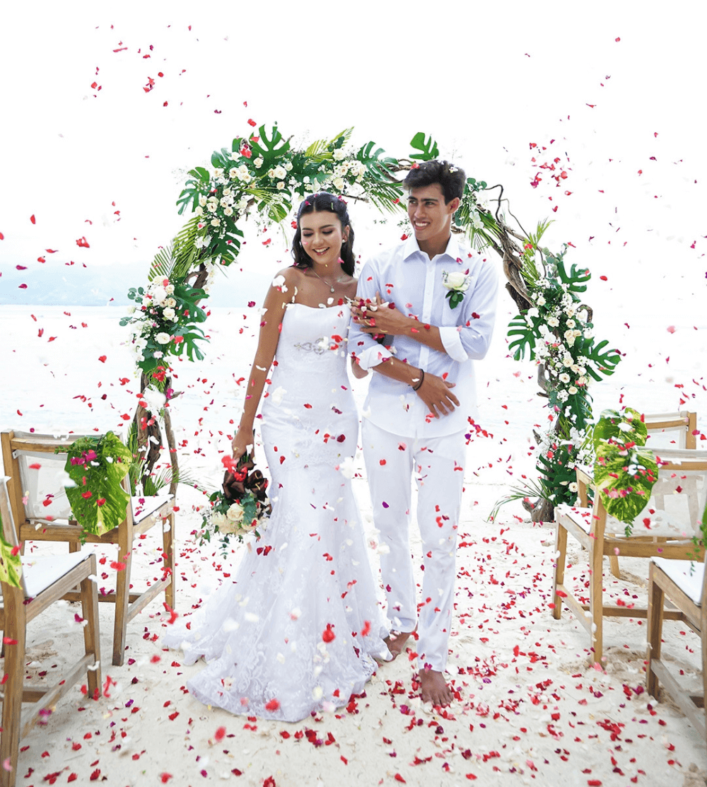 Newlywed couple at Pondok Santi Estate, Gili Trawangan, with wedding arch, blue waters, flowers in the air, and the bride holding flowers.